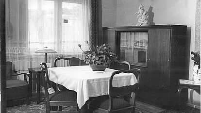 Blick in ein Wohnzimmer mit Tisch, Stühlen und Schrank, 1950er Jahre