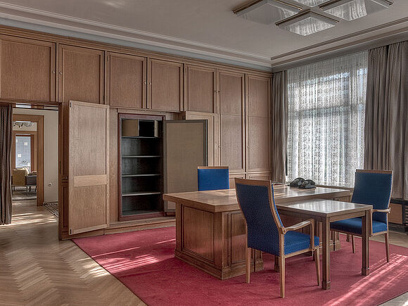 Das Bild zeigt ein hochwertig eingerichtetes Büro mit Holzverkleidung. In der Mitte des Raumes steht ein großer Schreibtsich mit drei Stühlen. Die Fenster sind mit halbdurchlässigen Vorhängen abgedeckt